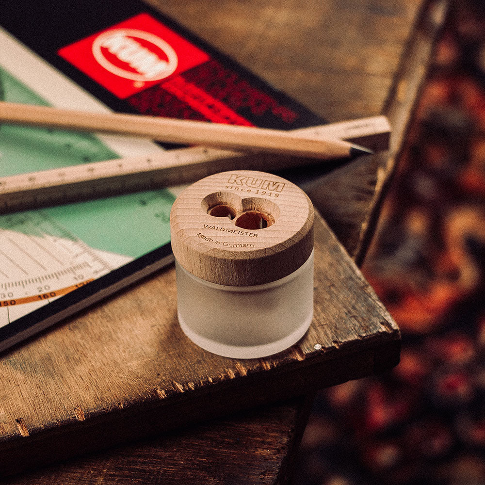 Small glass container with wooden lid on a wooden surface, with a ruler and other items in the background. KUM Waldmeister Sustainable Frosted Glass Pencil Sharpener Made in Germany