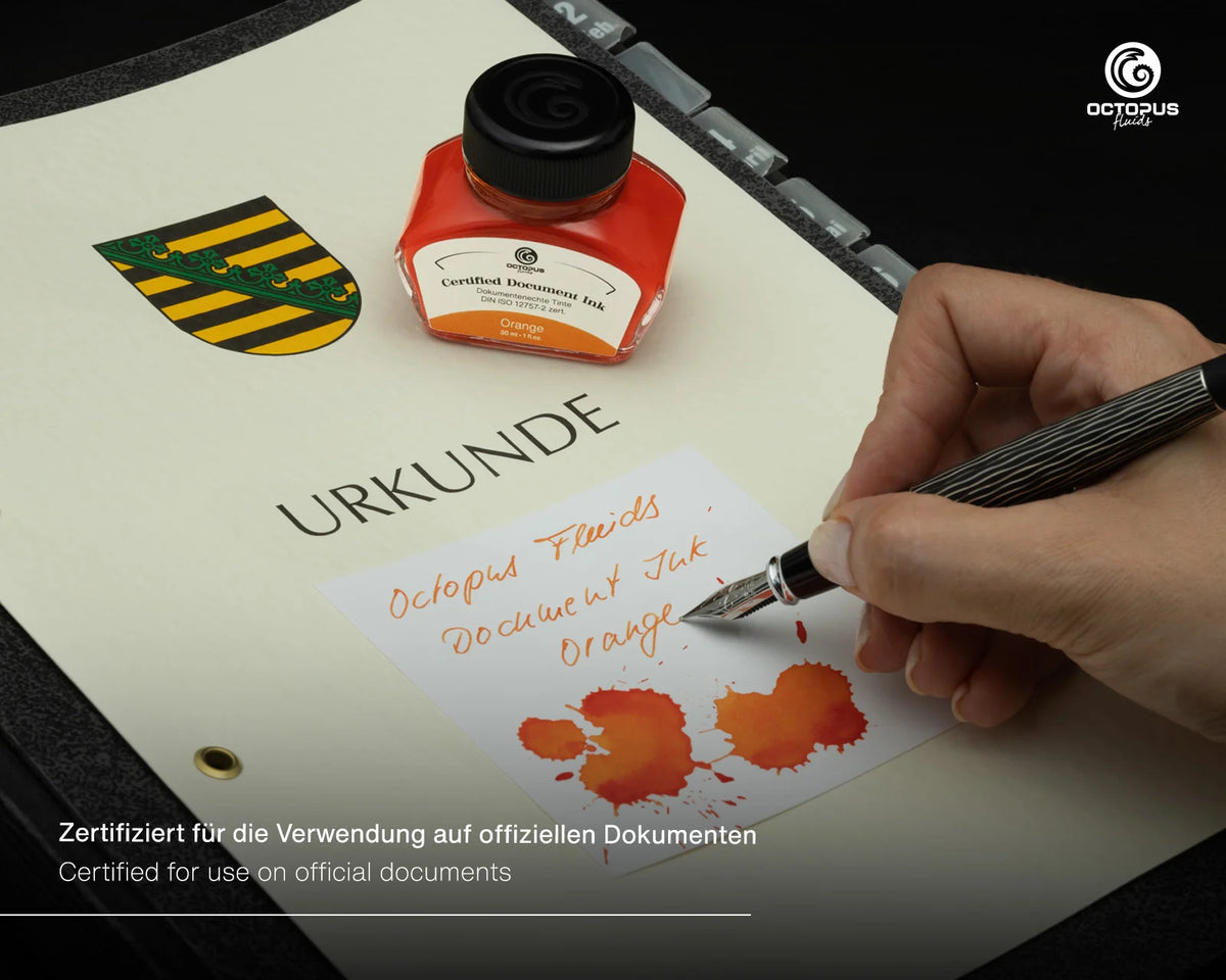 A hand holds a silver fountain pen over a white document, writing with orange ink. An octopus brand document ink bottle and a yellow-and-black crest are visible on the page, with certification text at the bottom. Octopus Fluids Fountain Pen Document Ink - 30 ml Bottle - Orange -  Made in Germany