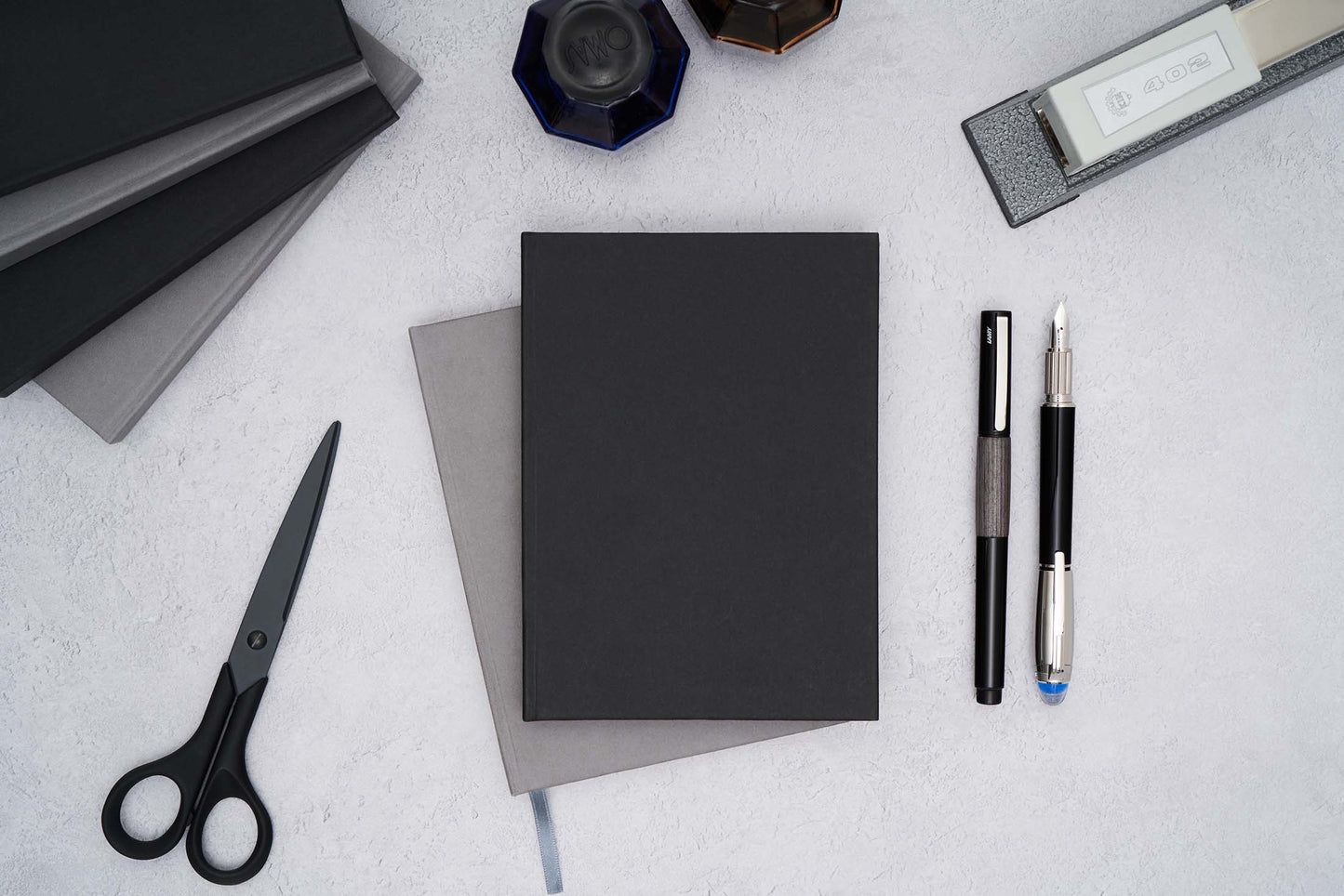 two the paper mind blocker paper notebooks stacked in gray and black on a flat surface with fountain pens ink a stapler and scissors 
