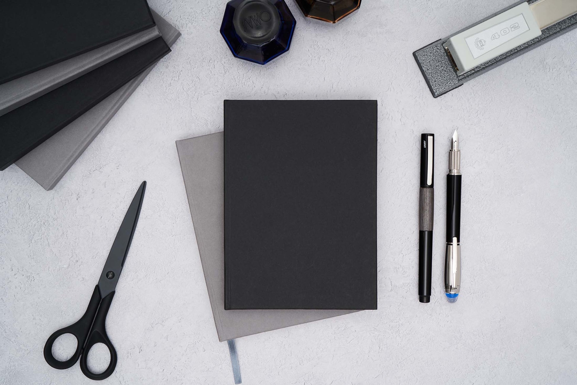 two the paper mind blocker paper notebooks stacked in gray and black on a flat surface with fountain pens ink a stapler and scissors 