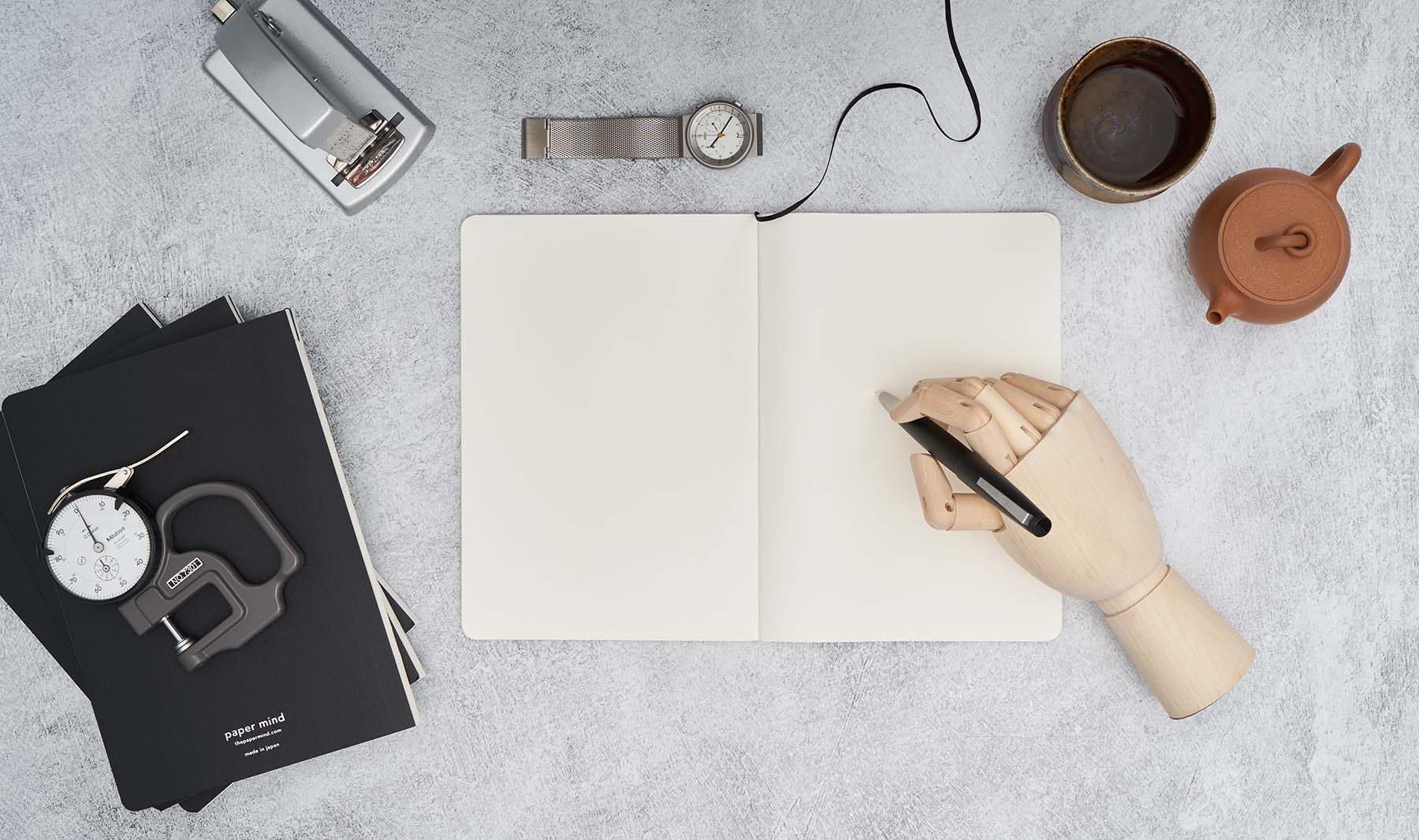 Paper Mind Mitsubishi Bank Paper Notebook open flat on gray background with wooden model hand holding Lamy 2000 fountain pen, zenith stapler, braun watch, yixing teapot on table